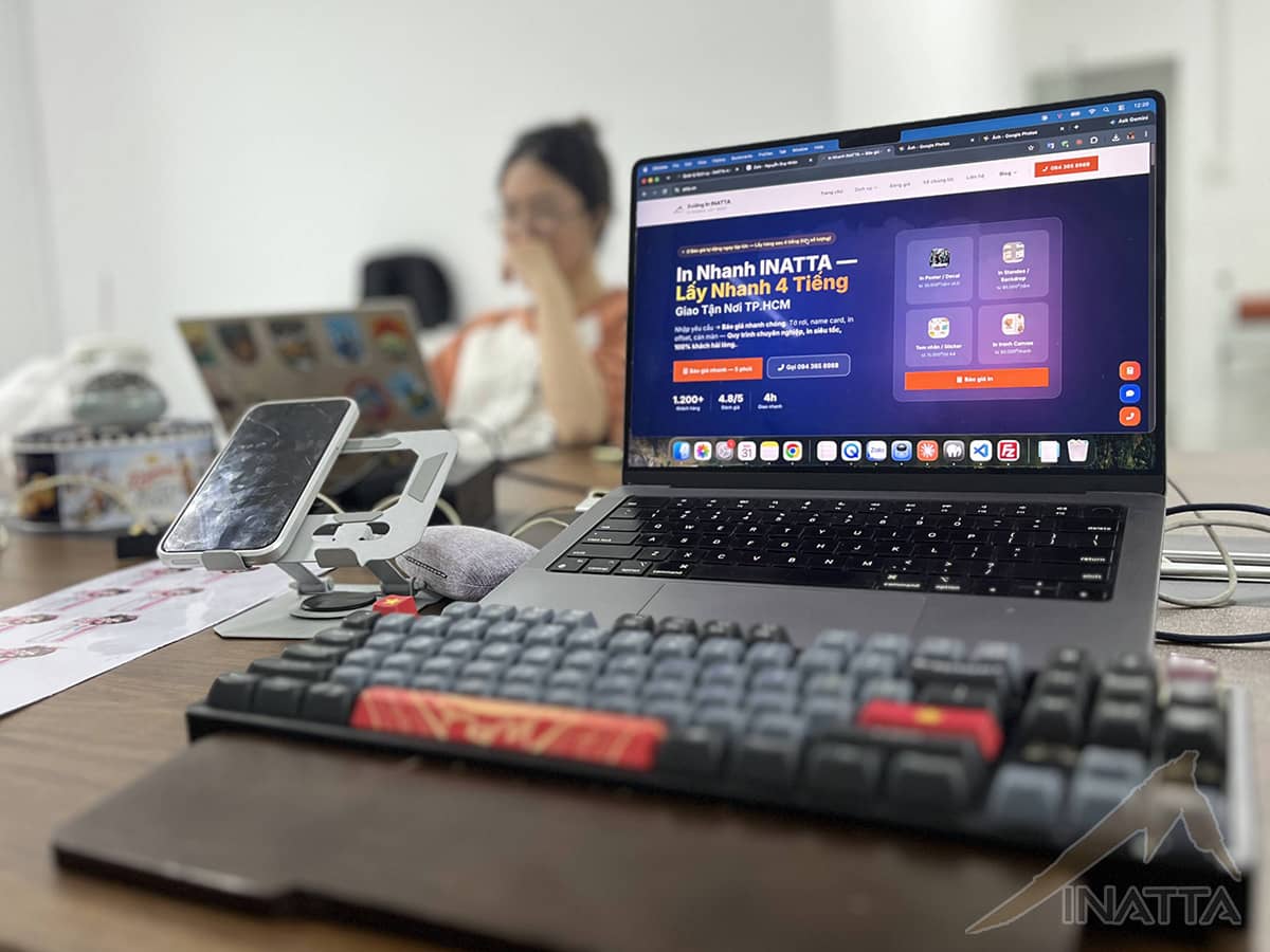 Góc văn phòng INATTA, màn hình laptop đang mở trang chủ atta.vn, nhân viên làm việc phía sau