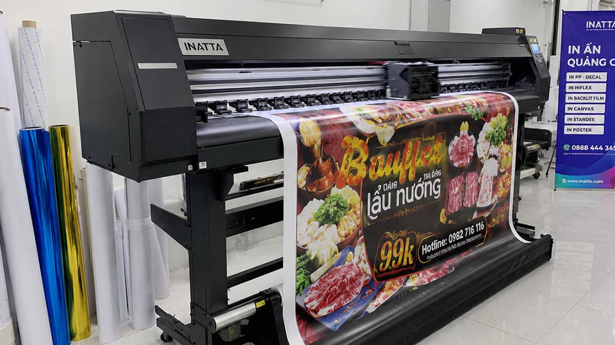 Ảnh chụp máy in khổ lớn trong xưởng in INATTA đang in poster trên chất liệu PP, đầu in chạy ngang trên cuộn vật liệu rộng, tấm poster màu vừa in ra được kéo xuống khay hứng, cuộn PP và decal dự trữ dựng bên cạnh, một standee đã hoàn thiện dựng phía sau, xưởng sạch đèn led trắng, chụp góc chéo lấy trọn thân máy, ảnh thương mại chân thực, tỉ lệ 4:3